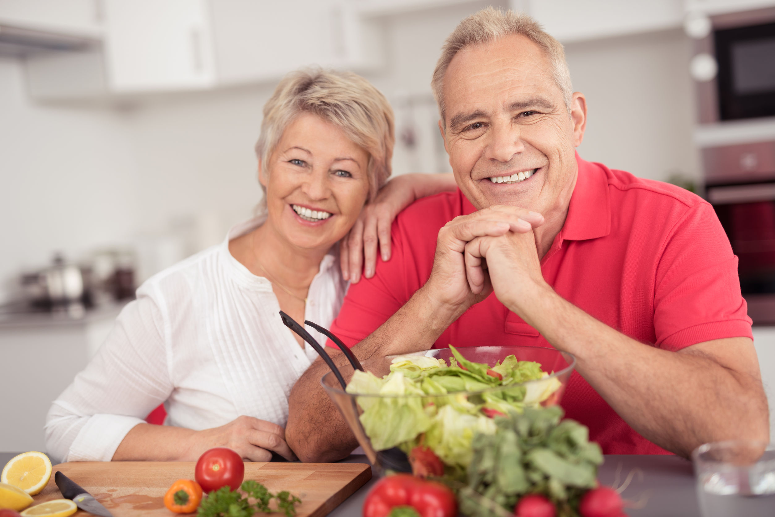 Portrait of a Happy Senior Couple Sitting at the Kitchen Table with a Bowl of Fresh Salad, Smiling at the Camera.