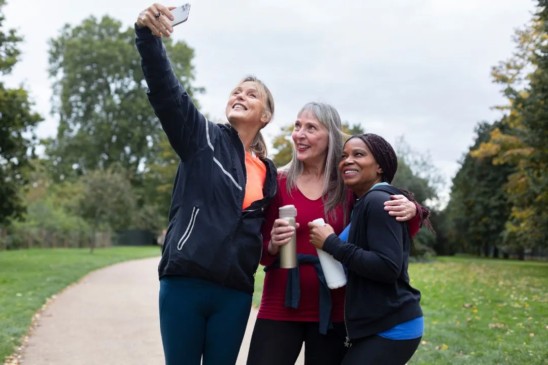 Three women smiling and posing for a selfie in a sunny park, surrounded by greenery and trees