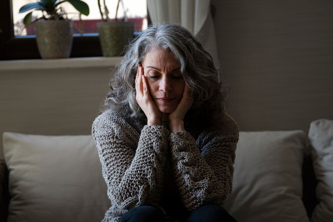 A woman sits on a couch, her hands covering her face, reflecting the weight of adult stress and emotional burden.