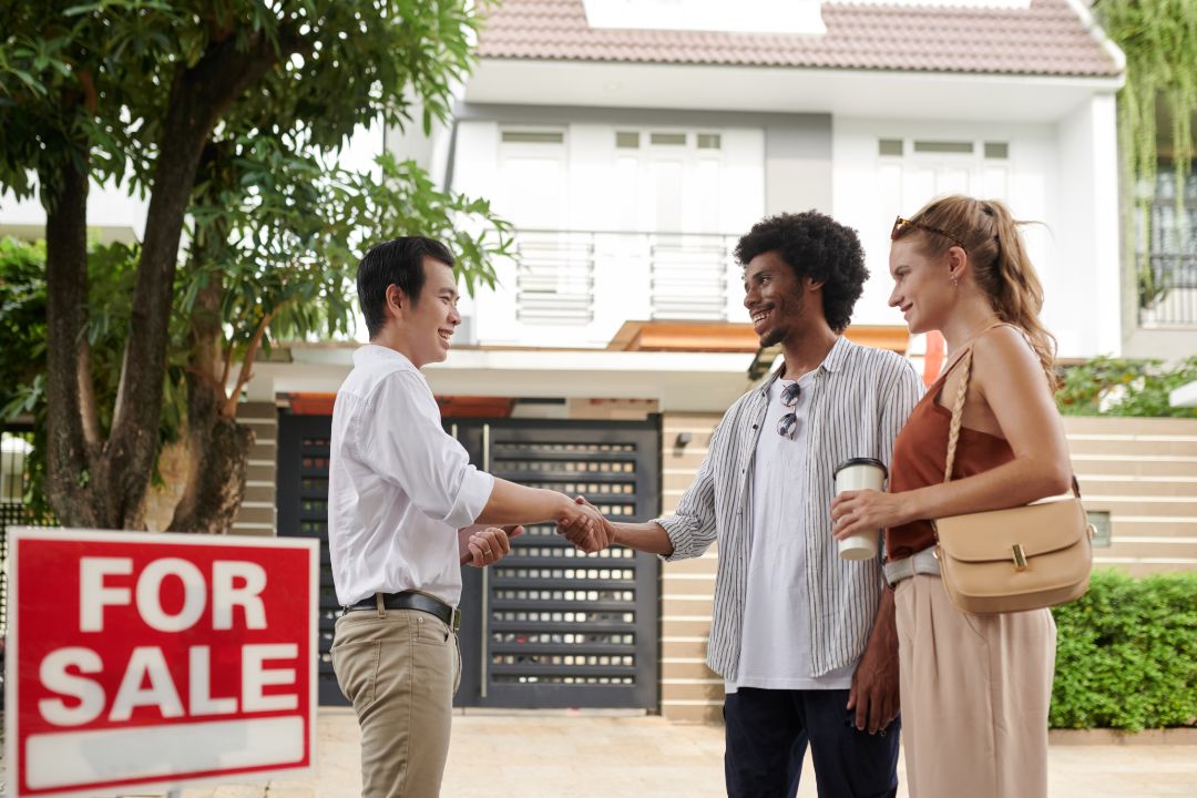 A couple engages in a handshake outside a house with a for sale sign, representing a positive agreement in real estate.