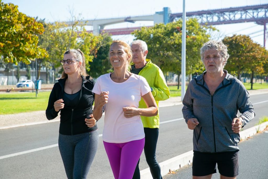 Four people, embracing senior fitness, jog on a sunny day with trees swaying gently in the breeze and a charming bridge in the background.