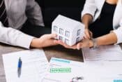 A couple proudly displays a paper house model and a pen, representing their recent mortgage approval.