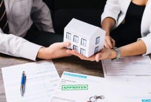 A couple proudly displays a paper house model and a pen, representing their recent mortgage approval.