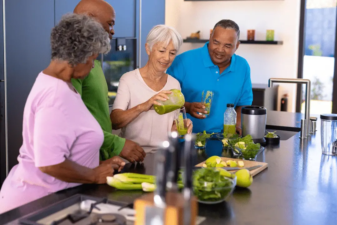 Four adults with an average age of 60 engaged in preparing healthy food in a kitchen . One woman with short gray hair and dark skin is pouring a green drink from a jar, while another woman with curly gray hair and light skin looks on. A man with light skin and a short head is standing next to them, and another man with dark skin is holding a drink . The kitchen has modern appliances and a countertop filled with fresh vegetables and fruits