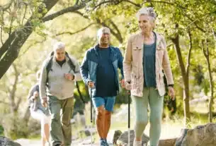 A group of older adults hiking on a forest trail, using walking poles. The trail is surrounded by green foliage and dappled sunlight.