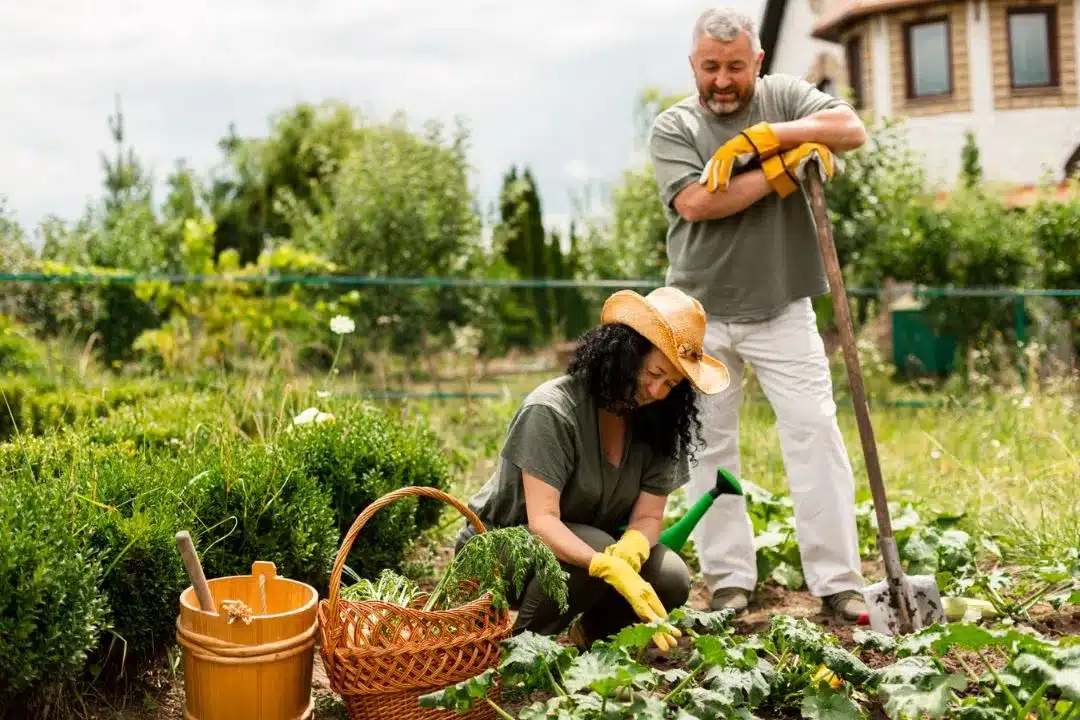 A woman in a straw hat and gardening gloves tends to plants in a lush garden while a man stands nearby with a spade.